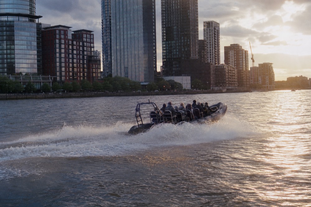 A twelve-seat rigid inflatable boat throwing a broad wake on the river in low golden light, city towers dark behind