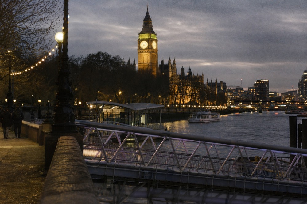 A river pier gangway at night lit by embankment lamps, the floodlit face of a Gothic clock tower rising behind the trees