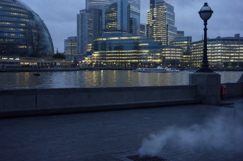 A wall of lit office towers seen low across black water at night, their reflections broken by a passing wake