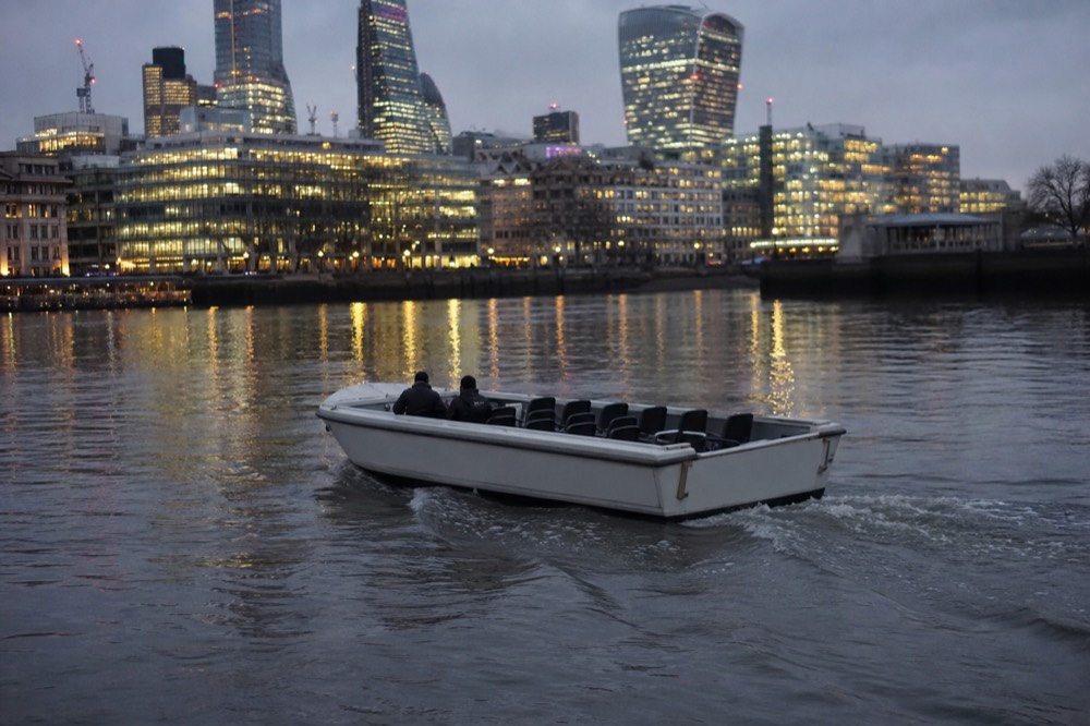 A small open charter boat crossing calm river water at dusk with lit buildings reflected behind, seats empty, no markings on the hull