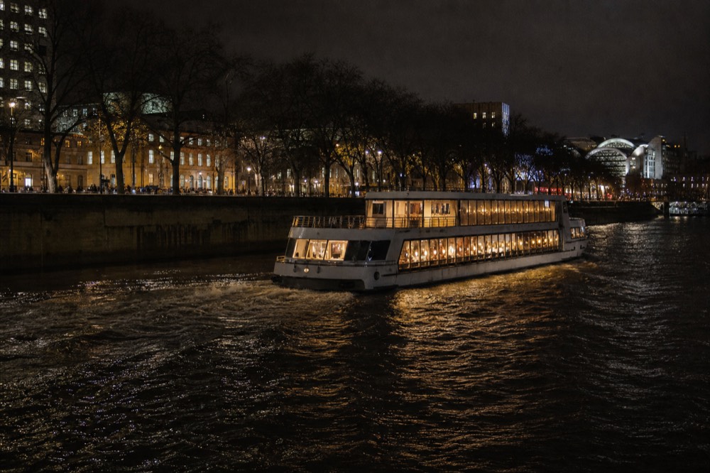 A larger river cruiser with warm lit windows passing a dark embankment at night, wake spreading behind it across black water