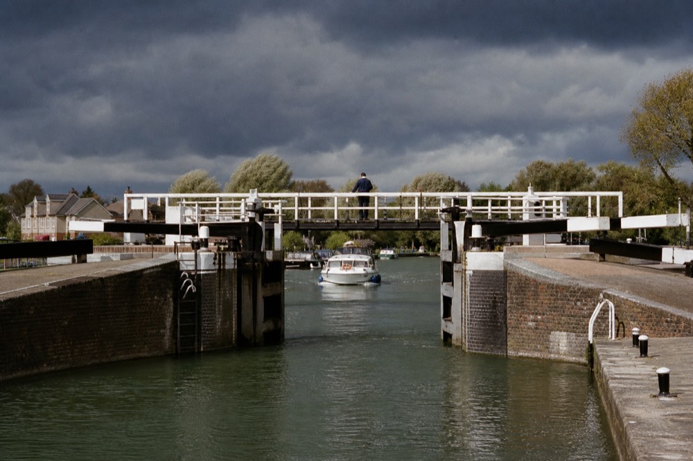 Open timber lock gates on a non-tidal river reach, green water held level between brick walls with a keeper's white footbridge above