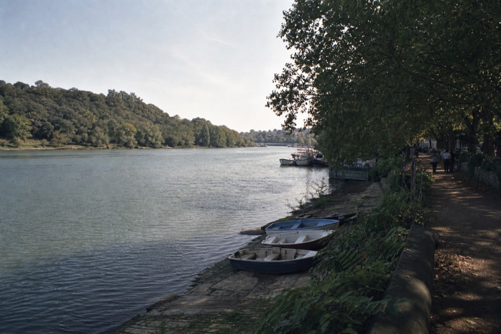 A wide green bend of river below a wooded hill on a bright afternoon, a towpath under mature trees and small boats drawn up on a slipway