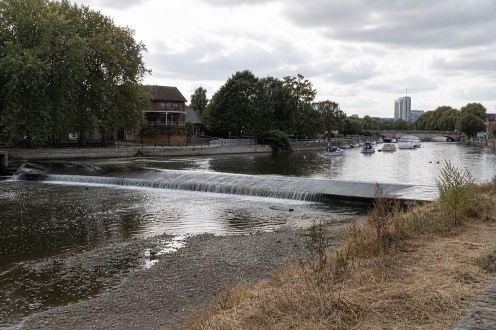 A low weir on a summer river with the water level noticeably down, exposed gravel shoals and dry grass on the bank in hard afternoon light