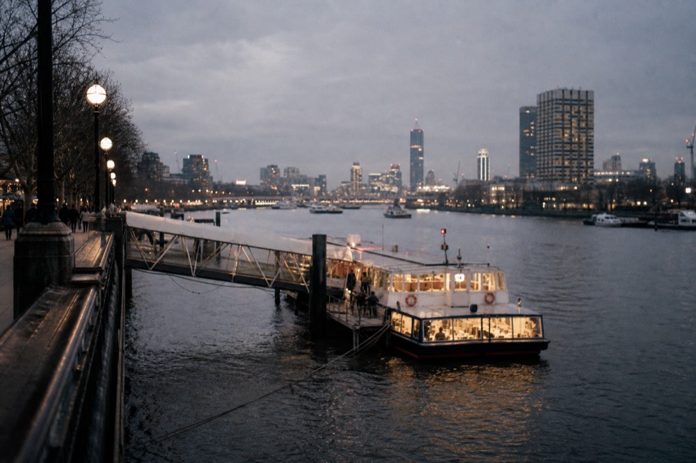 A floating pier at dusk with a gangway down to a lit passenger boat, the river dark and the embankment lamps just on
