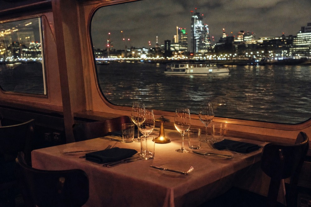 A laid table for four beside a boat window at night, wine glasses catching the light with the dark river and bank lights outside