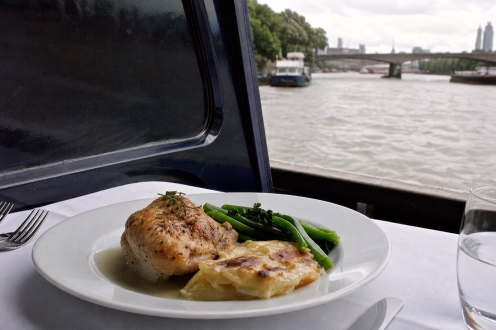 A plated chicken main course with dauphinoise potatoes and green vegetables on a white cloth, a boat window dark behind