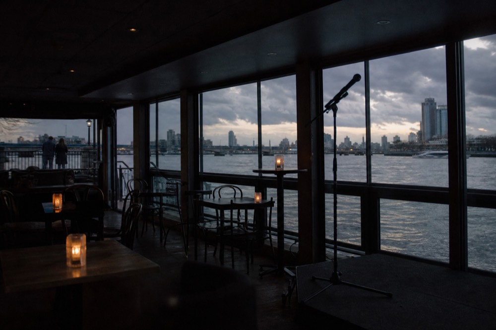 A dimly lit boat saloon in the evening with a microphone stand and small stage area, empty tables set with candles
