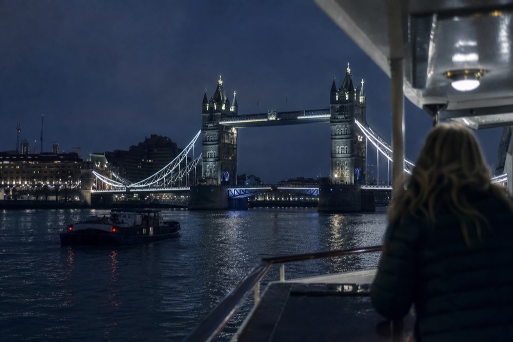 Tower Bridge floodlit from river level at night, seen over the rail of a passing boat with the water black and glossy