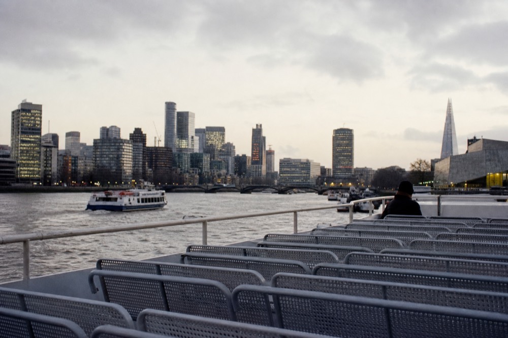 The open upper deck of a river cruise boat at midday with empty bench seating and a low rail, city towers behind