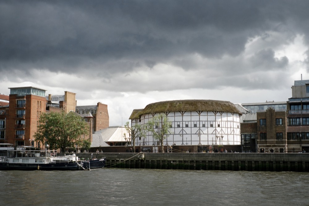 A round timber-framed theatre with a thatched roof on the south bank in bright daylight, brick warehouses to either side