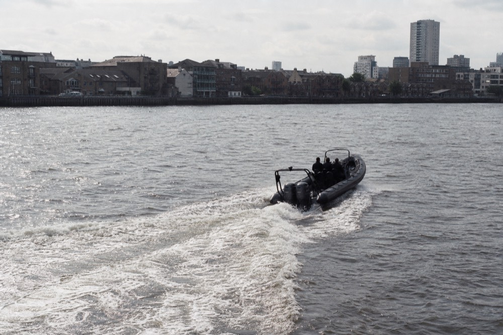 A rigid inflatable boat cutting a broad curving wake across open river water on a breezy afternoon, low wharf buildings on the far bank