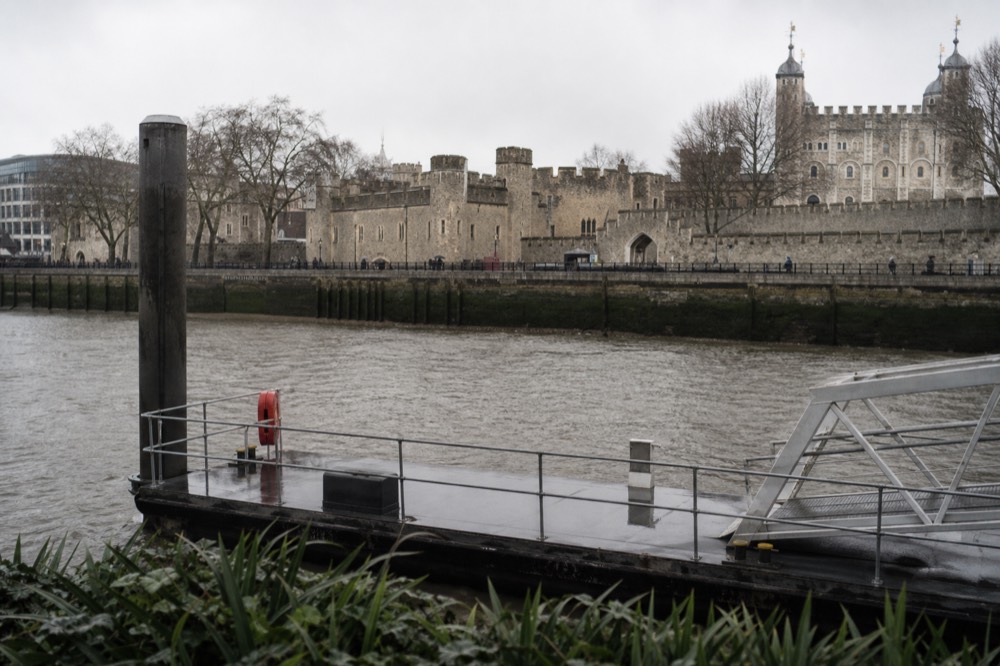 A floating pier pontoon beside the river on an overcast morning with the stone bulk of the Tower of London behind the embankment wall
