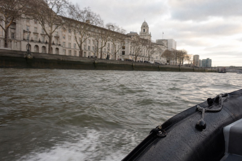 Choppy grey river water seen low from a small boat on an overcast day, spray on the tube of the hull, embankment wall in the distance