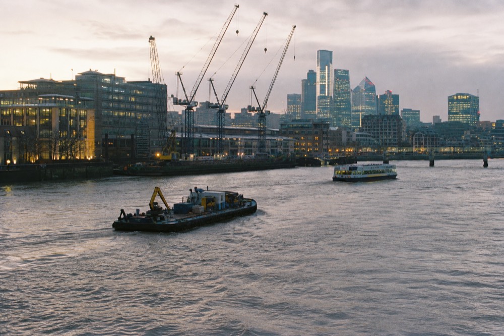 Mid-channel view of the tidal river with a working barge and a passenger boat passing on a clear day, wharf cranes on the south bank