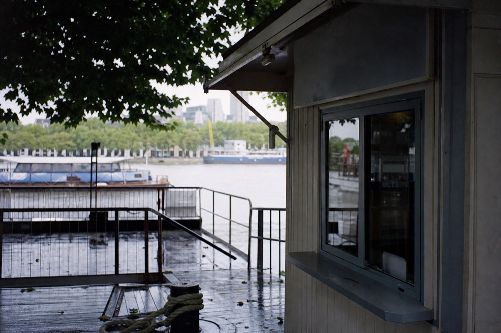 A small ticket window on a river pontoon with a price board above it, wet decking and a mooring rope in the foreground