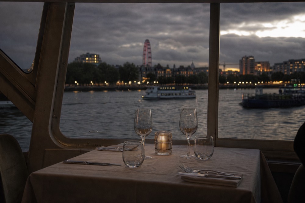 A laid table for four beside a boat window at dusk, cutlery and glasses set, the lit riverbank blurred outside