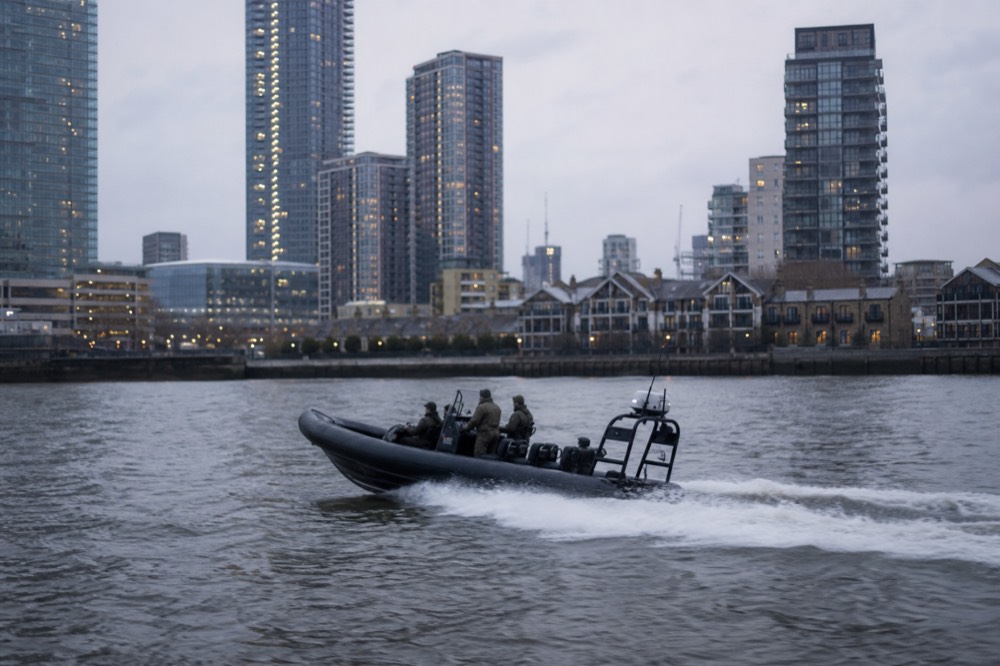 A twelve-seat rigid inflatable boat throwing a wide white wake on grey water with dockland towers rising behind
