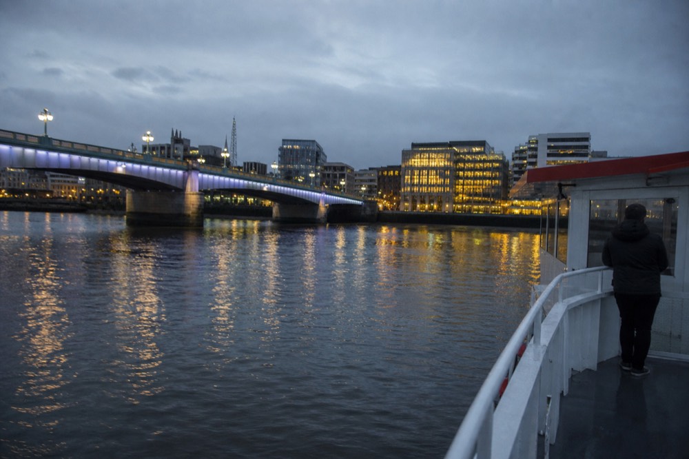 A floodlit bridge and lit riverside buildings reflected in flat dark water on a clear evening, a boat rail in the foreground