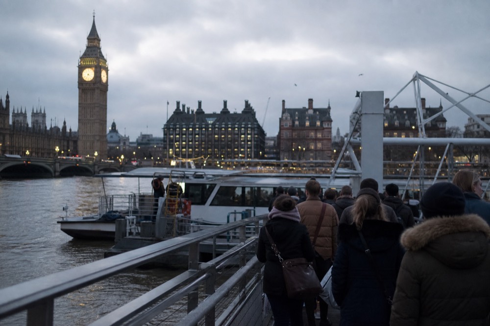 Passengers boarding a river cruise boat at a floating pier below a Gothic clock tower in early morning light