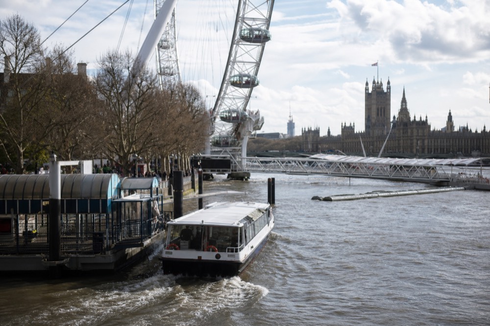A small sightseeing boat leaving a pier at the foot of a large observation wheel, riverside walkway busy behind