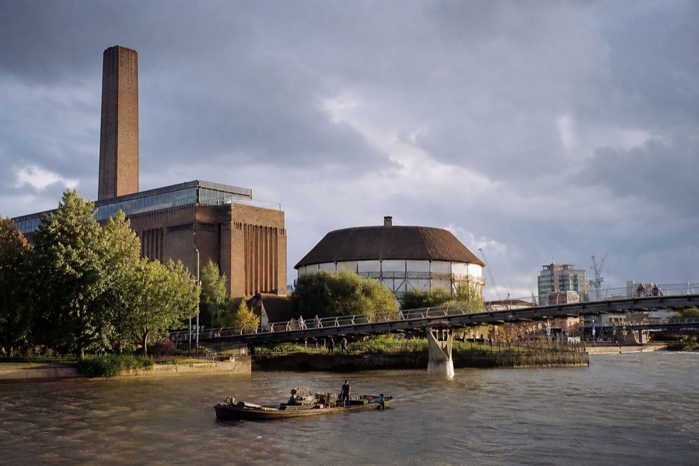 A brick former power station with a tall chimney beside a round thatched theatre on a river bank, footbridge crossing to the far side