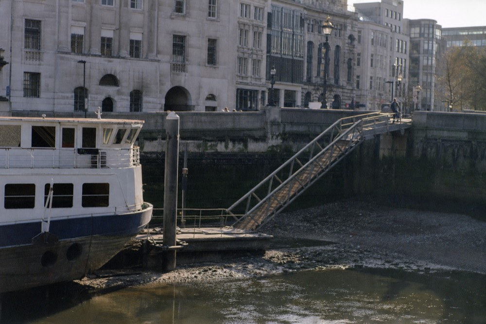 A floating pier pontoon with a steep hinged gangway at low water, a moored cruise boat and the embankment wall behind