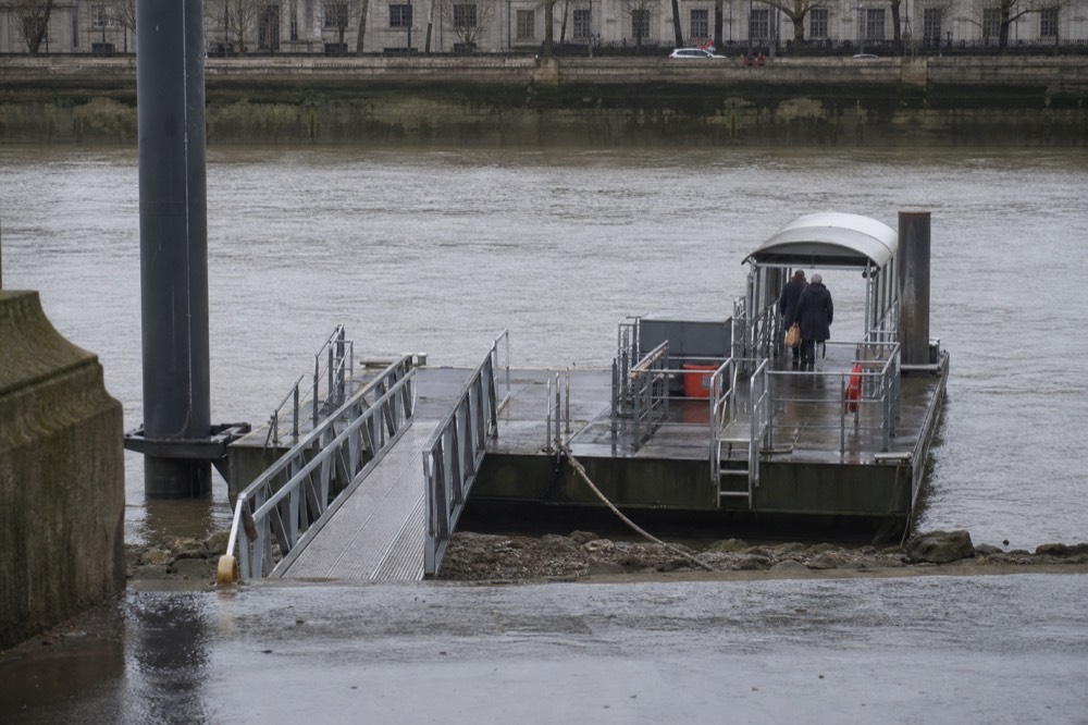 A floating pier pontoon with its gangway lowered on a bright morning, the river wide behind and the embankment wall above