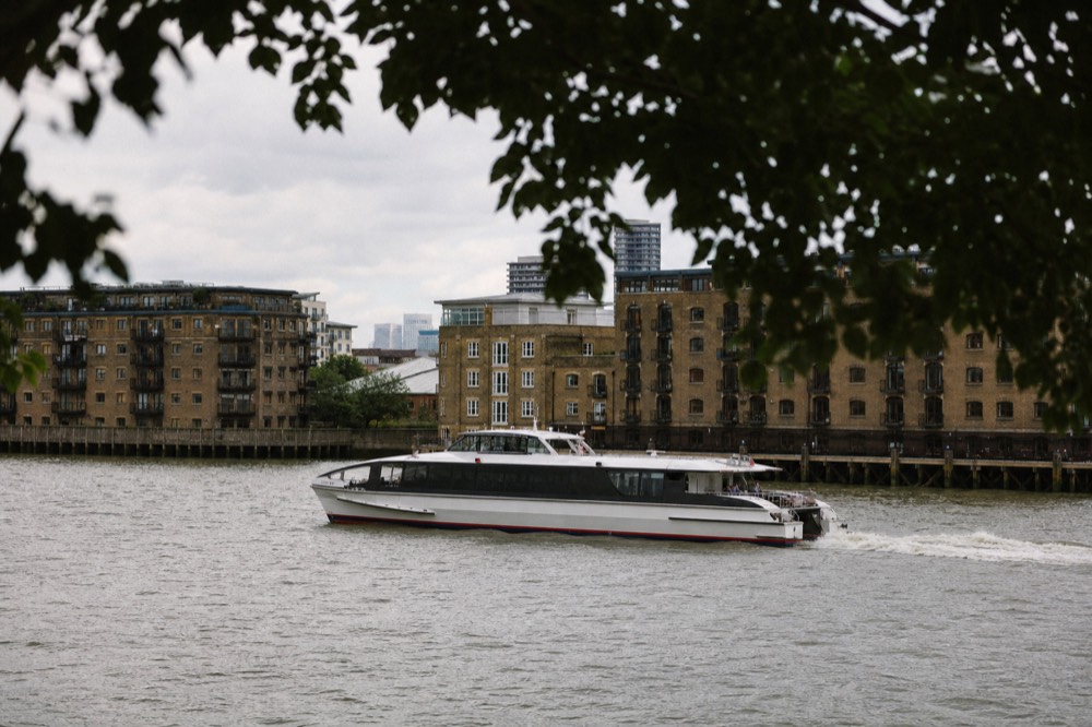 A fast twin-hulled passenger catamaran throwing a low wake on flat grey water with warehouse conversions along the bank behind