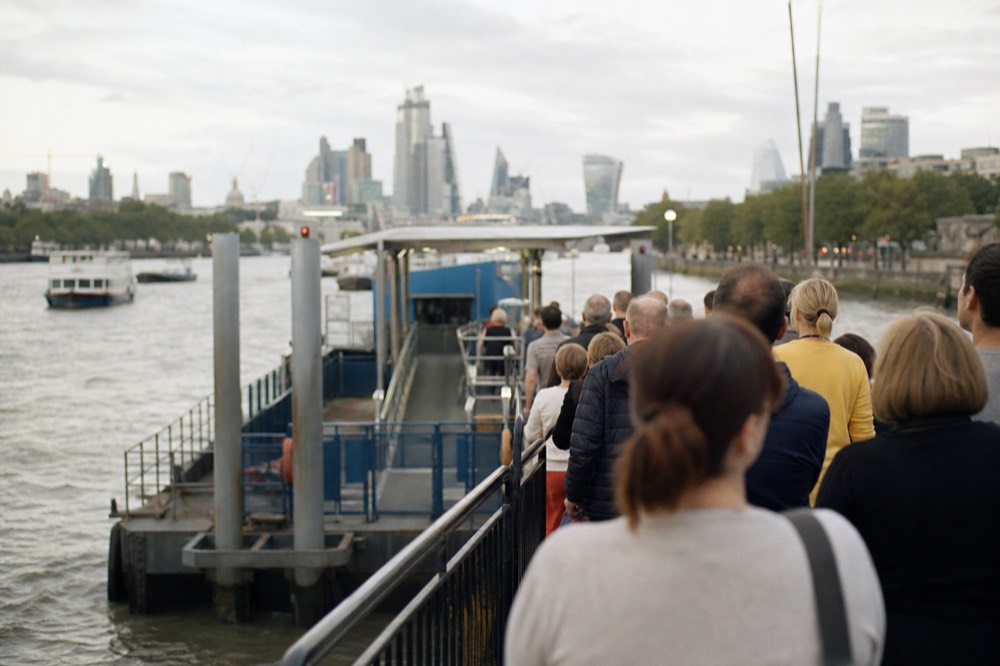 A line of people waiting behind a barrier at the entrance to a floating river pier on a bright summer morning, seen from behind