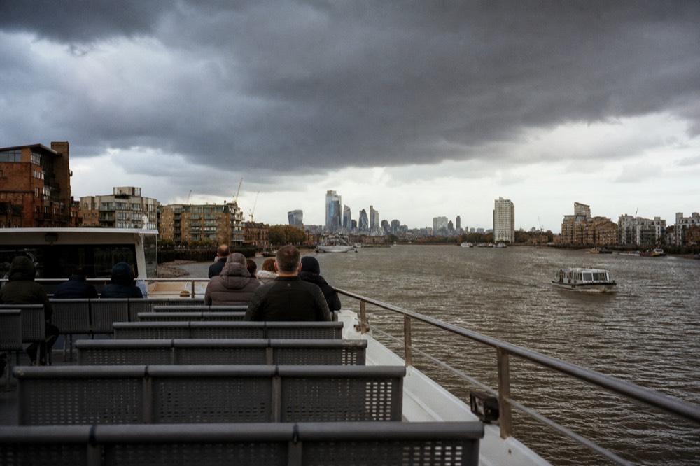 Seen from the back of an open upper deck, rows of empty bench seats and the river opening out towards a distant cluster of towers