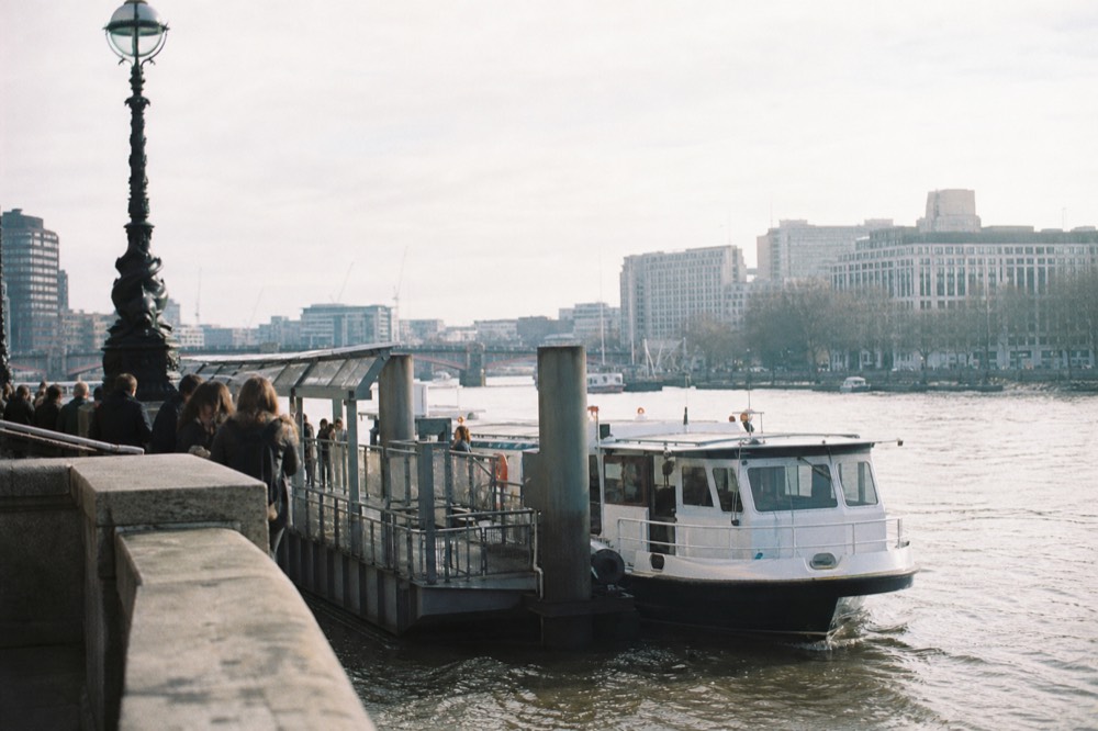 A floating pier beside a stone embankment on a bright morning, passengers waiting behind a rail as a white river boat comes alongside