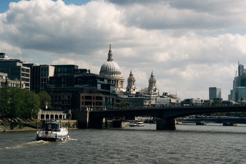 A pale stone cathedral dome above dark glass office blocks on a river's north bank, seen low from mid-stream under broken cloud