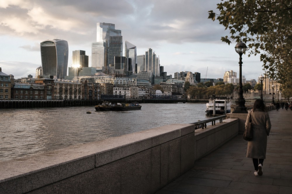 Tall glass towers of a financial district rising across a wide grey river reach, low wharf buildings along the near bank