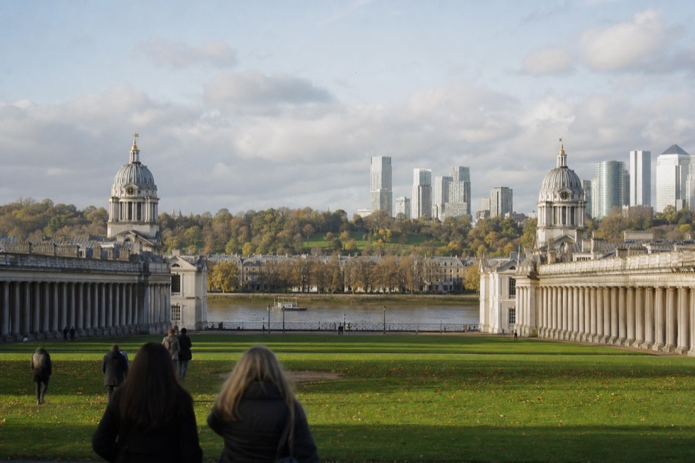 Two symmetrical colonnaded stone blocks either side of a wide lawn running down to a river, a green park hill rising behind