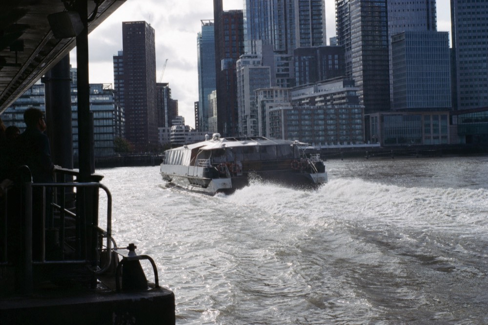 A fast catamaran river bus throwing spray as it accelerates away from a pier, city towers grey behind on a bright cold day